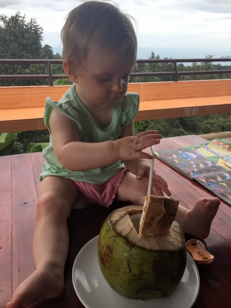 Our youngest child enjoying fresh coconut water while seated at an outdoor wooden table in Thailand, a perfect highlight for a family itinerary with kids.