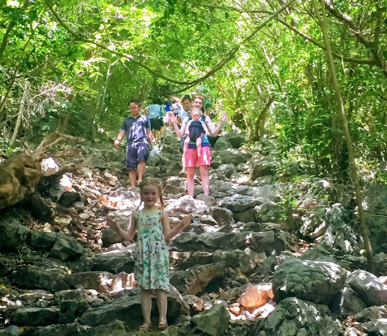 Hikers walking up a rocky trail surrounded by dense greenery, heading towards the iconic Phraya Nakhon Cave in Thailand.
