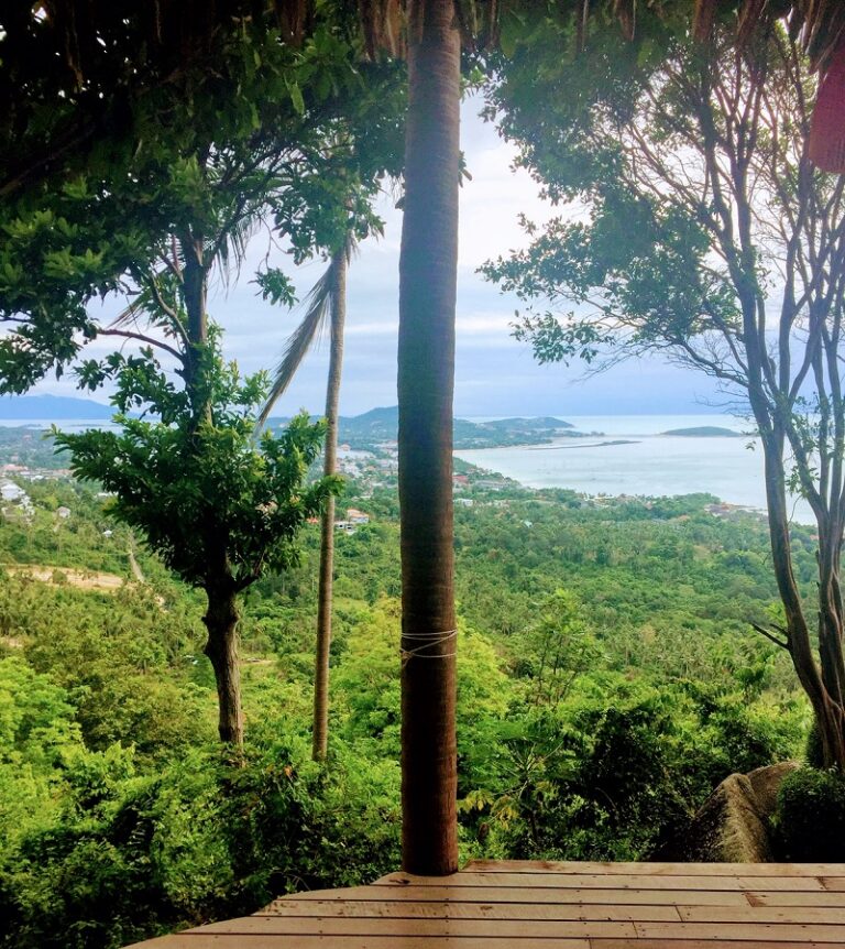 Scenic view of Koh Samui, Thailand, featuring a wooden deck, tropical greenery, and the coastline with the sea extending to the horizon.