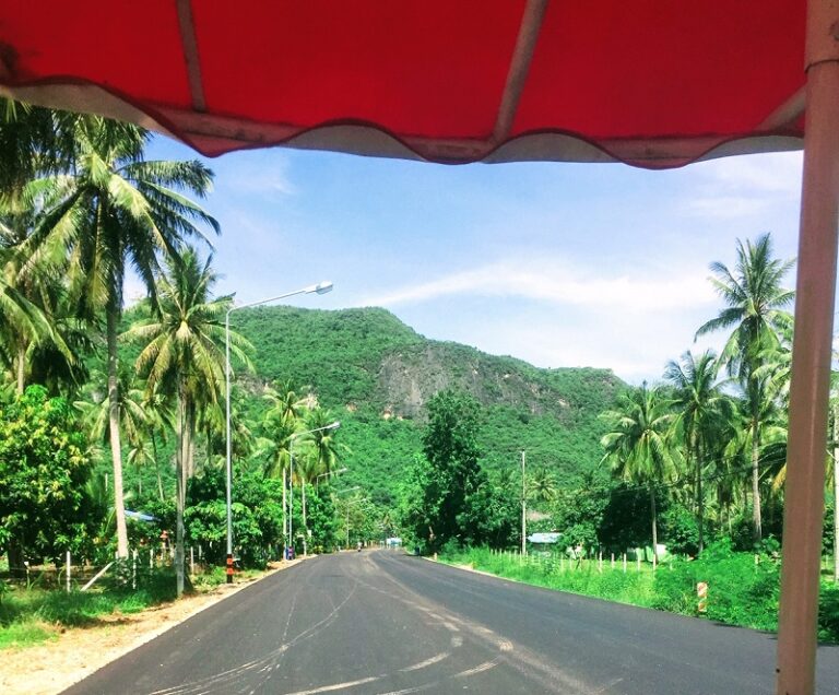 Scenic road in Sam Roi Yot, Thailand, flanked by palm trees and tropical vegetation, with green mountains in the background.