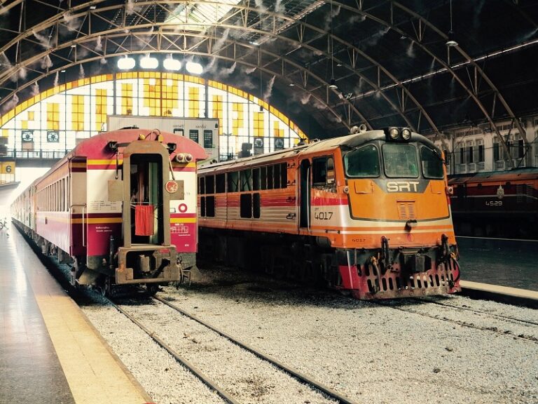 Two trains at Hua Lamphong Station in Bangkok, Thailand, featuring a red-and-cream passenger train and an orange locomotive under a stained glass arched roof.
