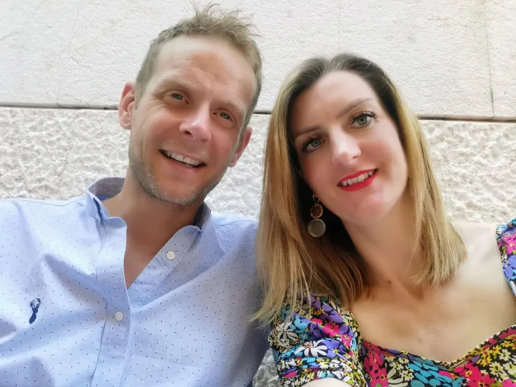 Me and Lee seated side by side—Lee wearing a light blue polka-dot shirt with an embroidered logo, and myself in a colorful floral dress. A textured wall with a horizontal dividing line serves as the backdrop.