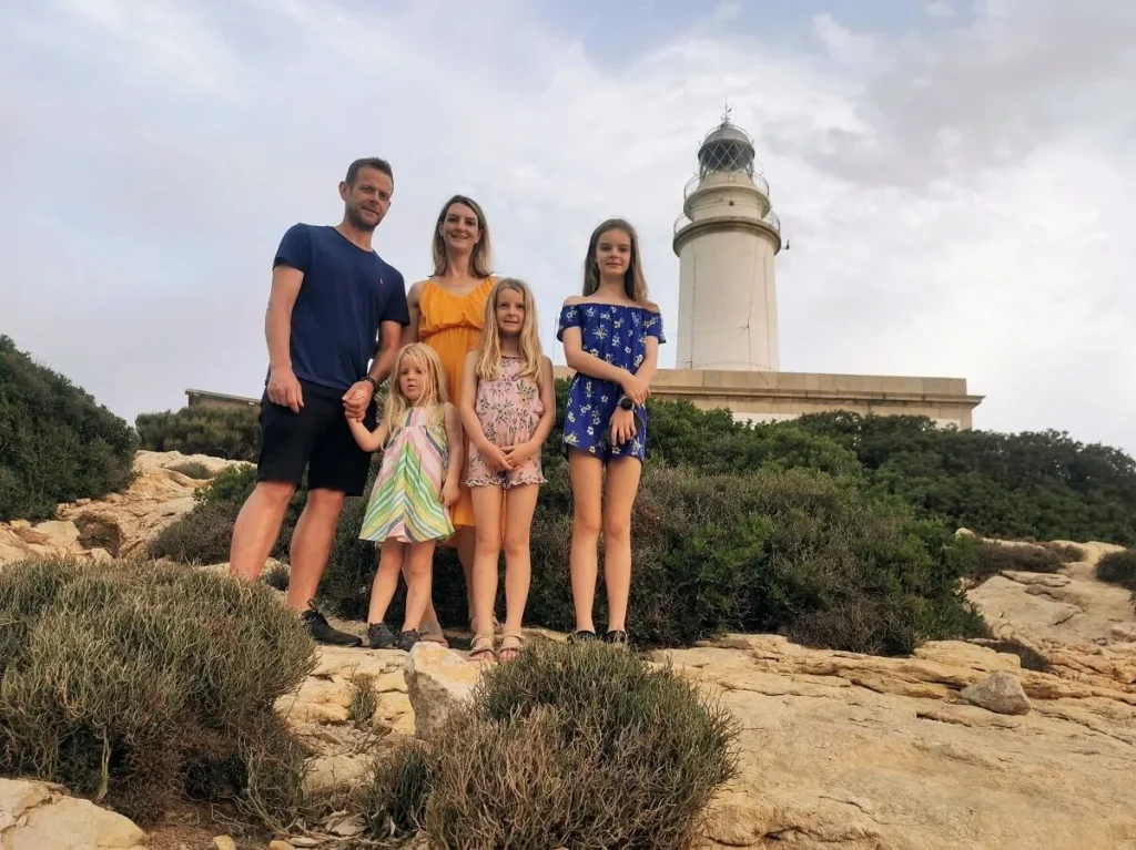 Myself (Mum), Lee (Dad) and our three kids, standing on rocky ground with the Formentor lighthouse in the background. We stand on either side of the children, adding to the sense of togetherness and adventure.