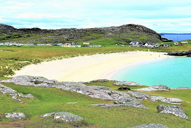Clear turquoise water and sandy shores of Achmelvich Beach with surrounding hills and cliffs under a bright sky.