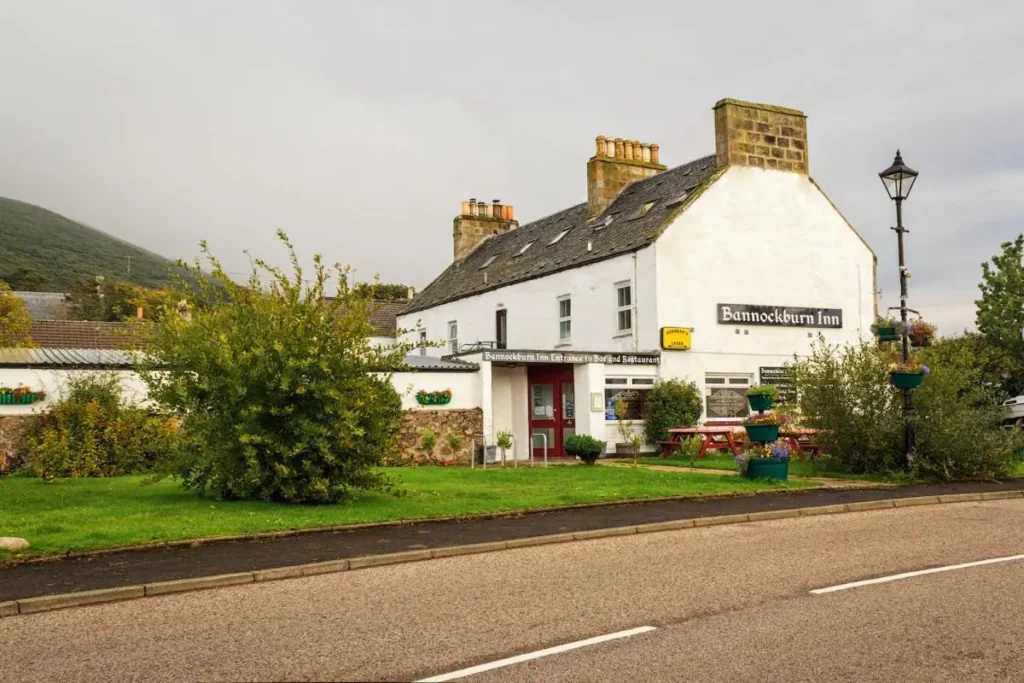 A different angle of Bannockburn Inn, a white corner building with signage for bar and accommodation services. The entrance is framed by a stop sign, street lamp, and flower arrangements. The inn’s outdoor seating area features wooden picnic tables surrounded by greenery.