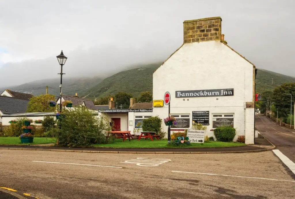 A view of Bannockburn Inn, a white building with signage, located at a street corner with a backdrop of misty hills. The inn has outdoor seating and a garden area with flowers and shrubs.