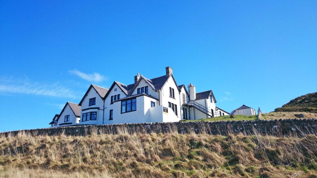 The front view of Bettyhill Hotel, a traditional Scottish NC500 hotel with a white exterior, set against the backdrop of Torrisdale Bay. The entrance features signage and a welcoming atmosphere.