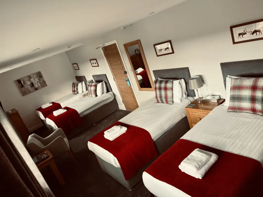 A well-appointed quad room at Bettyhill Hotel, featuring four neatly arranged single beds, wooden furnishings, and soft lighting. The space includes a window with curtains, offering a view of the surroundings.