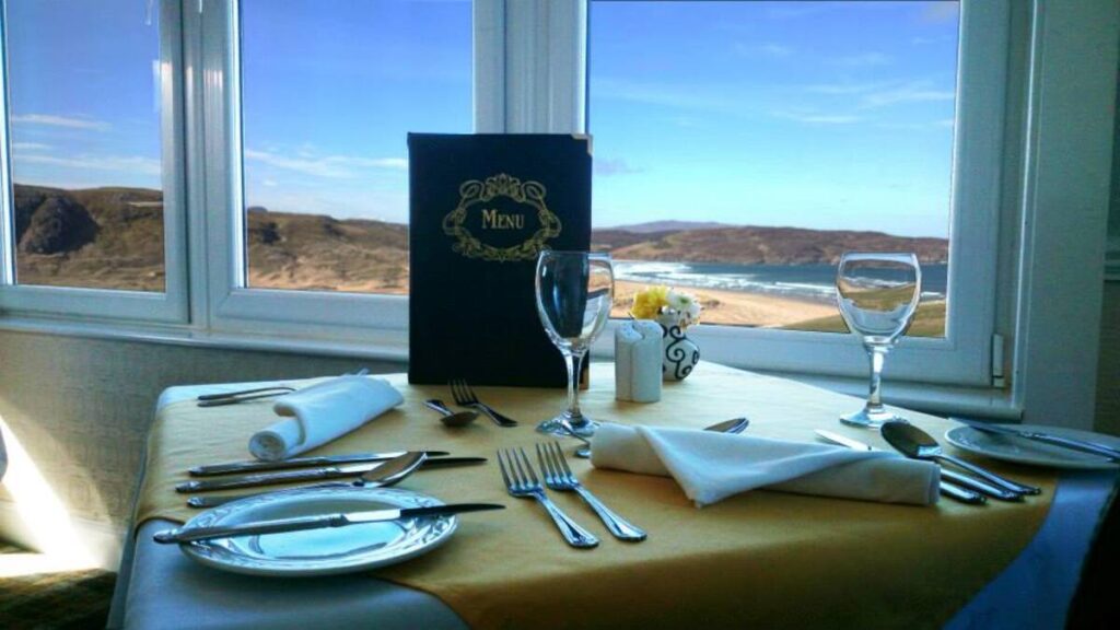 The Bettyhill Hotel restaurant, featuring a cozy dining space with wooden tables, chairs, and large windows offering scenic views of Torrisdale Bay. Soft lighting enhances the inviting atmosphere.