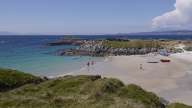Expansive sandy beach of Camusdarach, with calm turquoise waters and surrounding hills, under a clear sky.