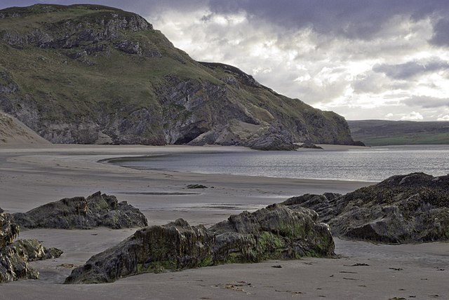 View of Coldbackie Beach in evening light, with golden sand, distant hills, and a calm sea under a soft sky.