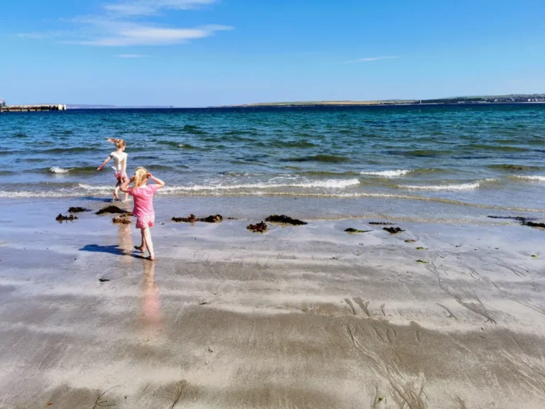 A sandy beach with two children playing near the water, small waves crashing onto the shore, and a pier extending into the ocean under a mostly clear sky.