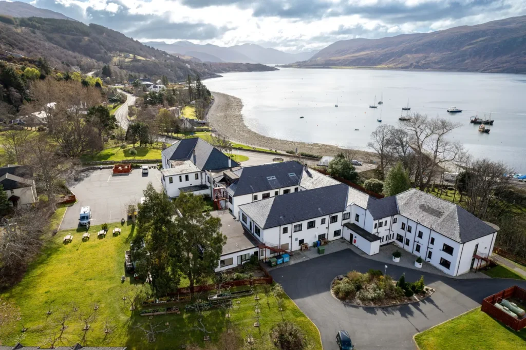 A bird’s-eye view of a seaside hotel in Ullapool, Scotland, surrounded by streets and parked cars, with water in the background. The hotel features a red roof and white exterior, standing near the waterfront.