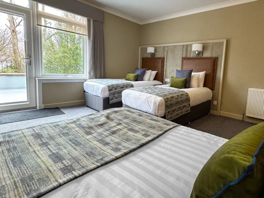 A cozy hotel room in Ullapool, Scotland, featuring a neatly made double bed with plush pillows, wooden furniture, and soft lighting. The space includes a window with curtains, offering a glimpse of the outdoors.