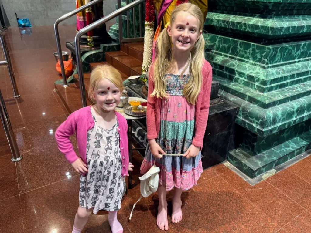Our two children in pink cardigans standing barefoot near offerings inside a Malaysian temple during a family-friendly cultural stop.