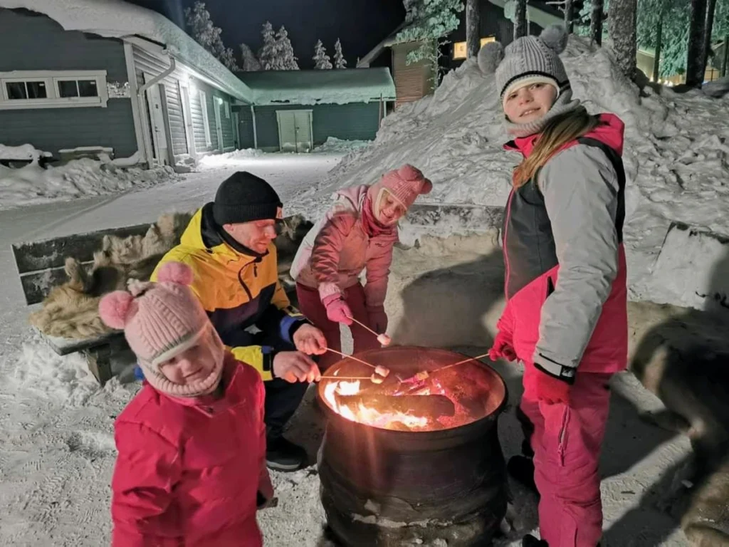 Our family roasting marshmallows over a fire pit in snowy Lapland, outside a kota-style hut.